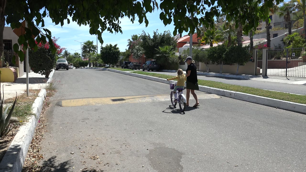 Older woman stands beside a child on a bicycle in a quiet street.