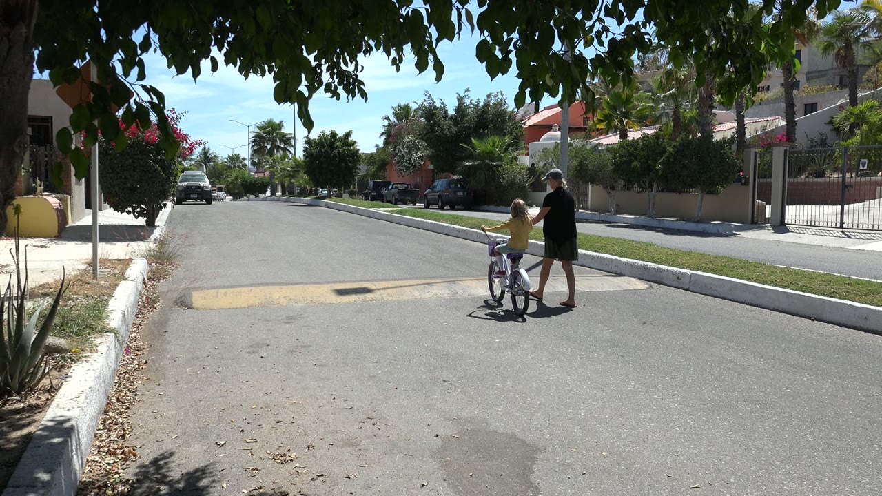 One second in, the older woman walks at the child's side as the bicycle is positioned near the center of the Los Cabos residential street, with tree shade across the top of frame.