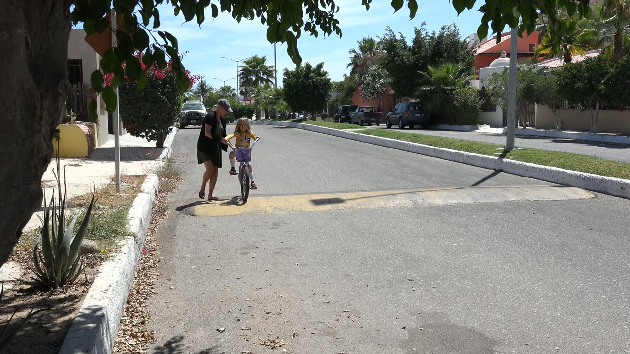 In Los Cabos, Mexico, an adult woman stands beside a young girl on a small bicycle near a shaded curb at the start of the clip.