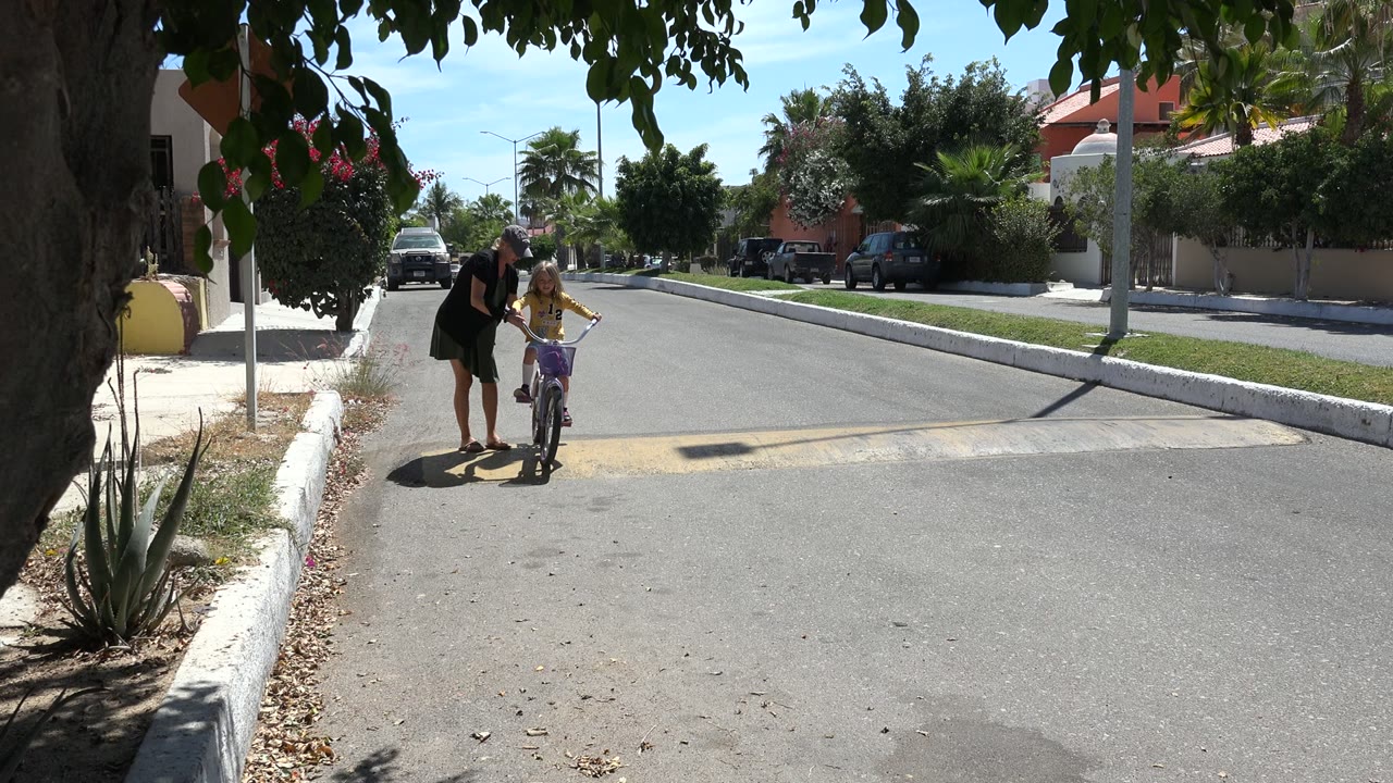 The adult woman leans toward the young girl as the child balances the bicycle near the curb on a sunlit Los Cabos street.
