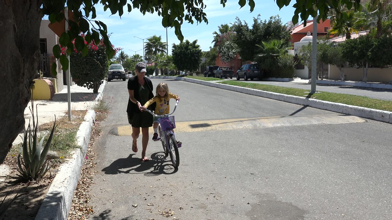 Adult woman helps a young girl on a bicycle in a sunny street.