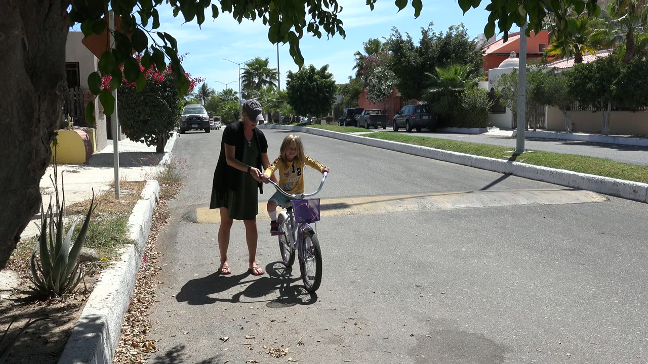 One second in, the woman keeps one hand near the bicycle while the girl faces forward on the residential street in Los Cabos.