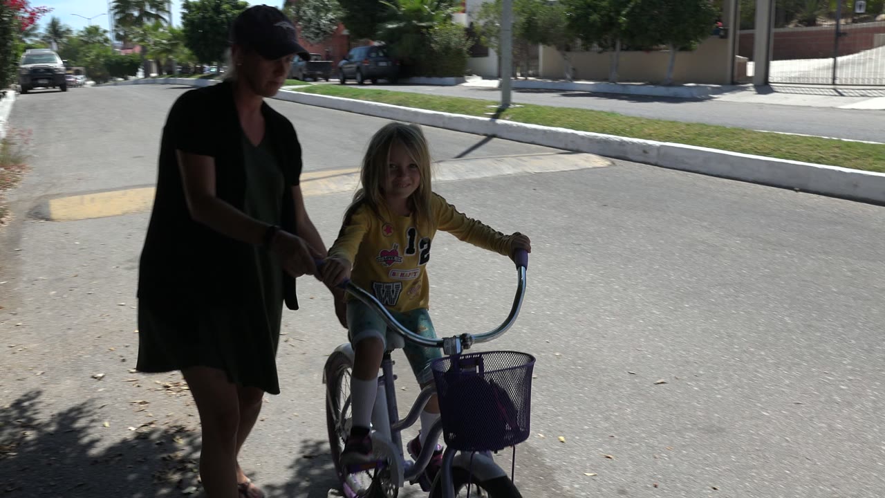 Adult woman helps a child on a small bicycle in a shaded street.