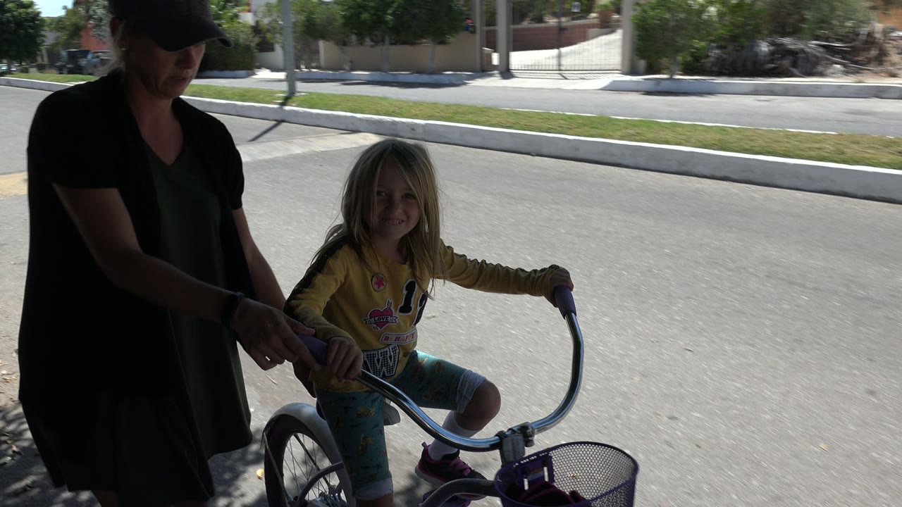 In Los Cabos, Mexico, the child smiles on the bicycle while the adult woman keeps one hand near the seat or handle area, with bright pavement and a residential wall behind them.