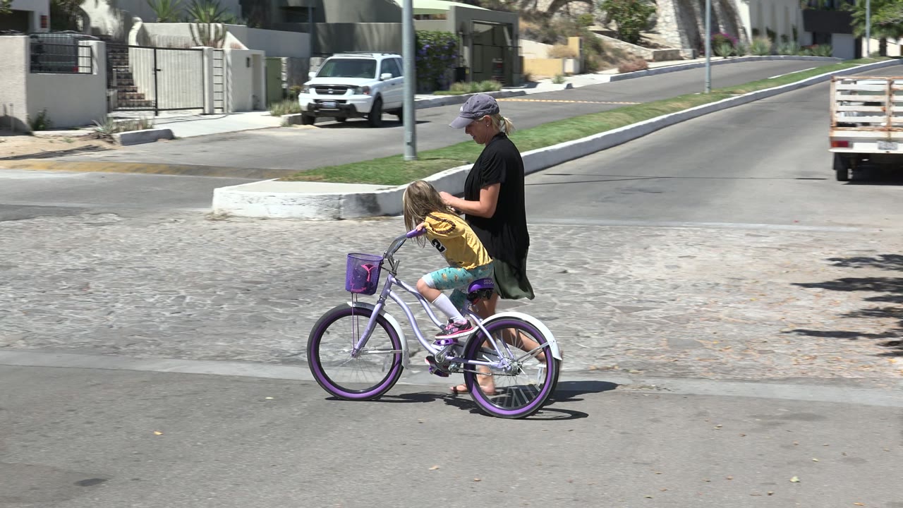 Adult guiding a child on a purple bicycle in a sunny street.