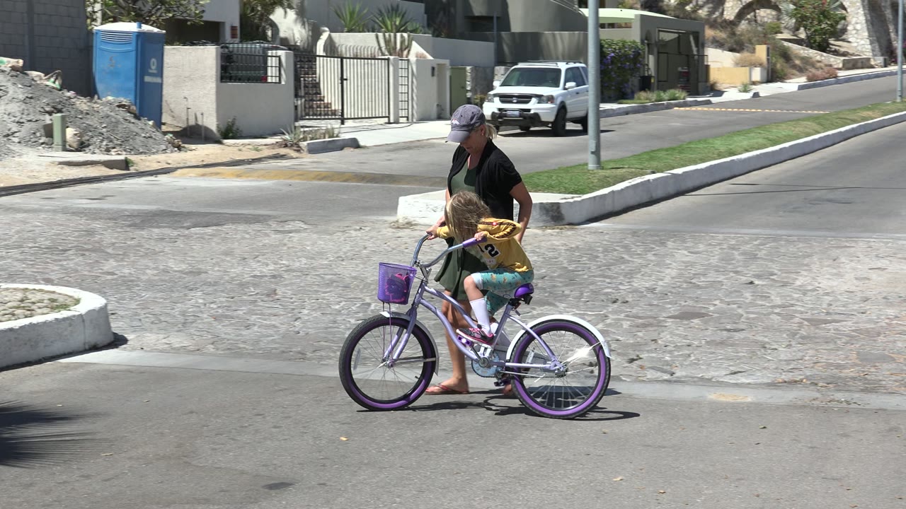 The child leans forward on the purple bicycle as her father stays close beside the seat, with gravel piles, a portable toilet, a gated home, and a parked white vehicle behind them in Los Cabos, Mexico.