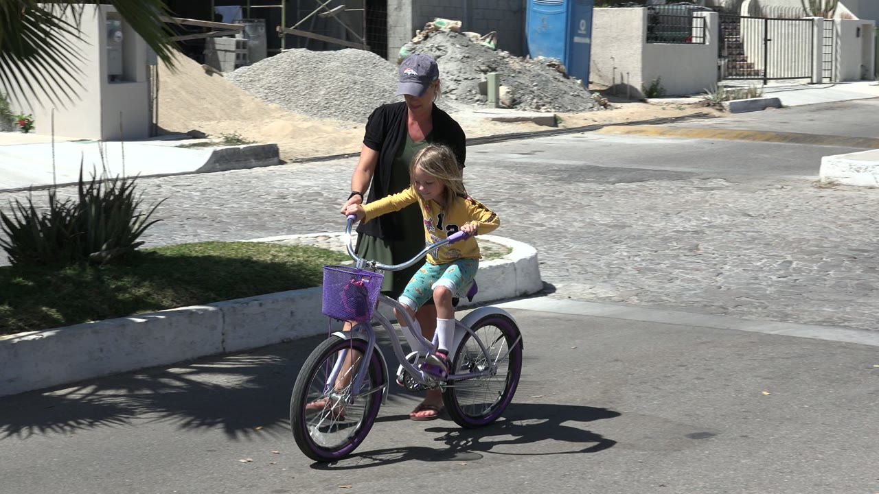 Adult woman guiding a young girl on a bicycle beside a sunny curb.