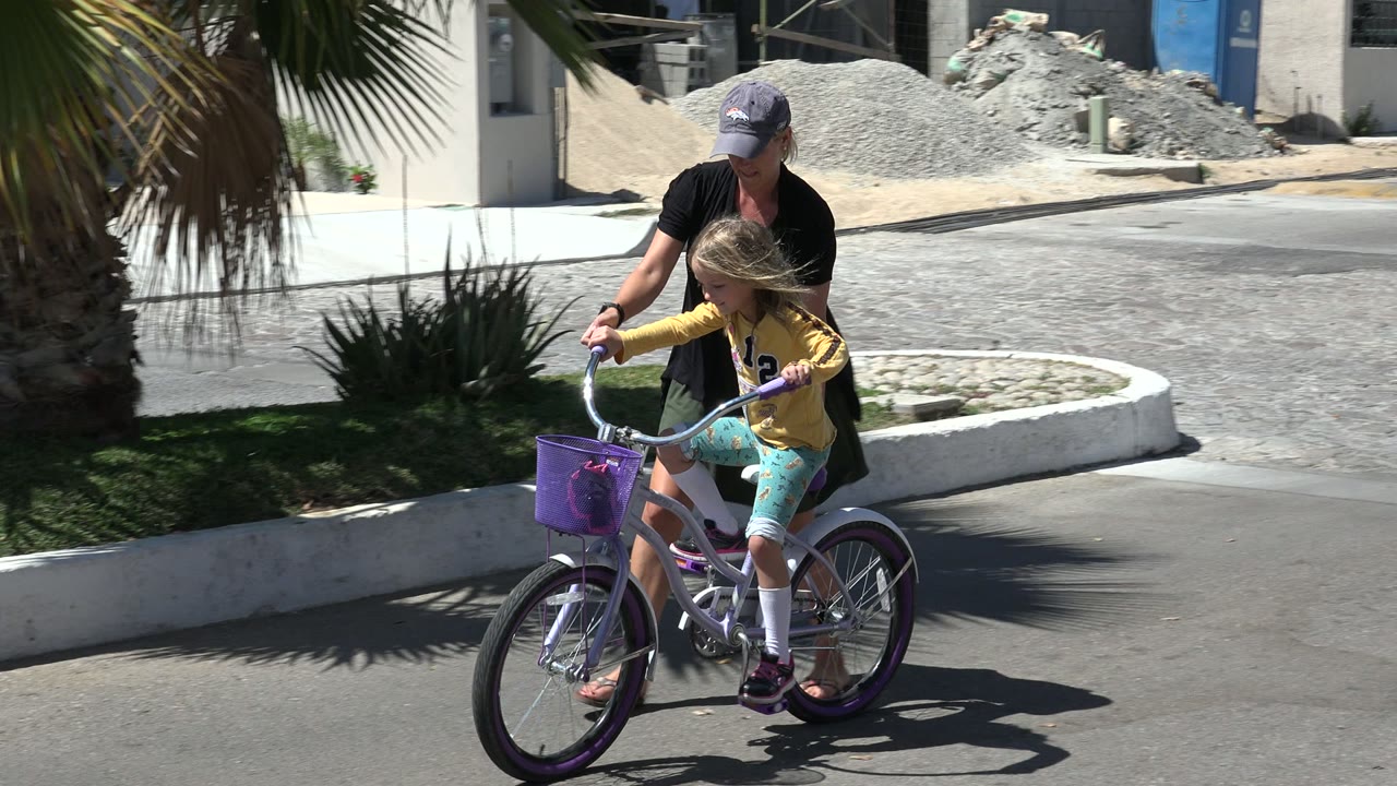 The child pedals forward on the small bicycle while the adult woman remains just behind the seat on a bright residential street in Los Cabos, Mexico.