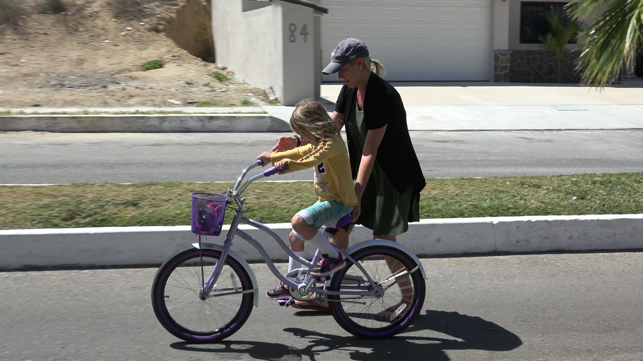 Adult woman walking behind a child riding a purple bicycle on a sunny street.