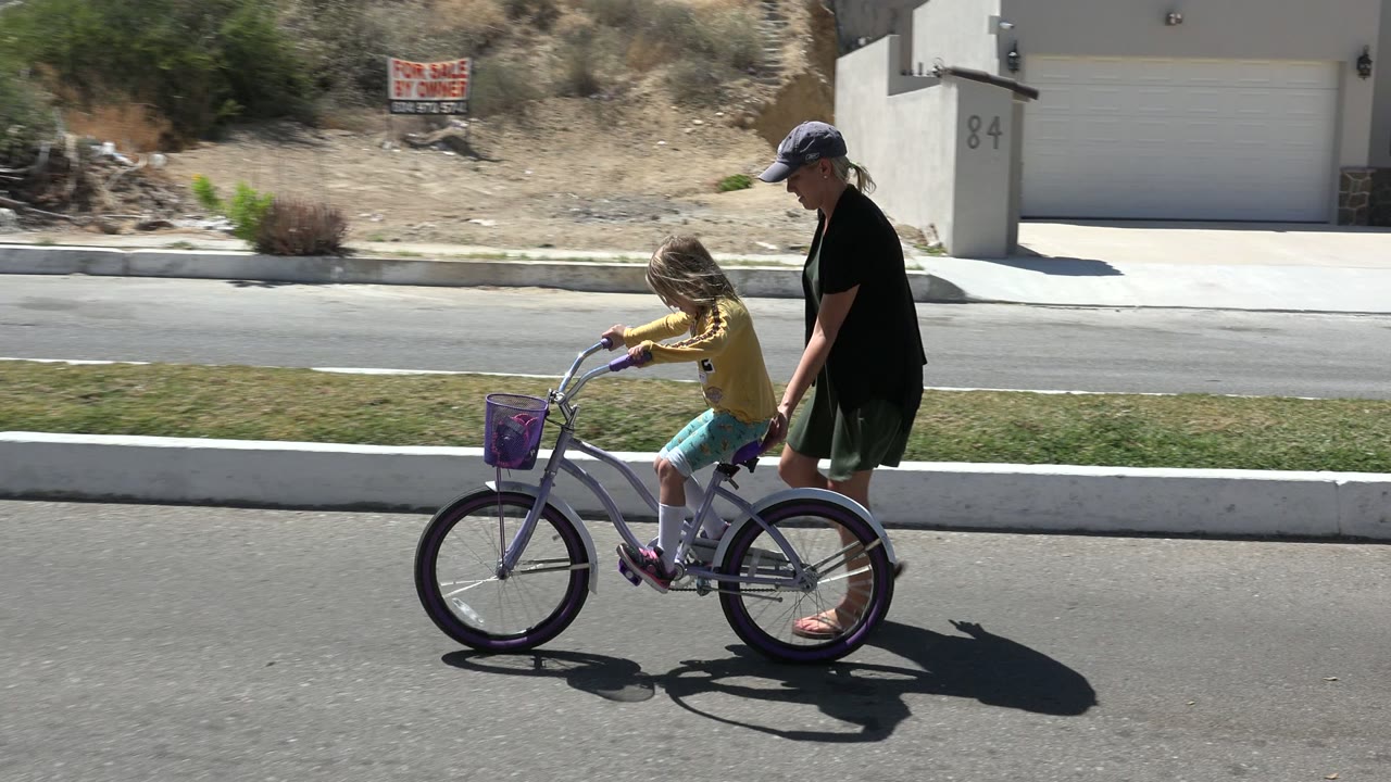 In Los Cabos, Mexico, the child pedals forward on the purple bicycle as the adult woman follows just behind on the sunlit road.