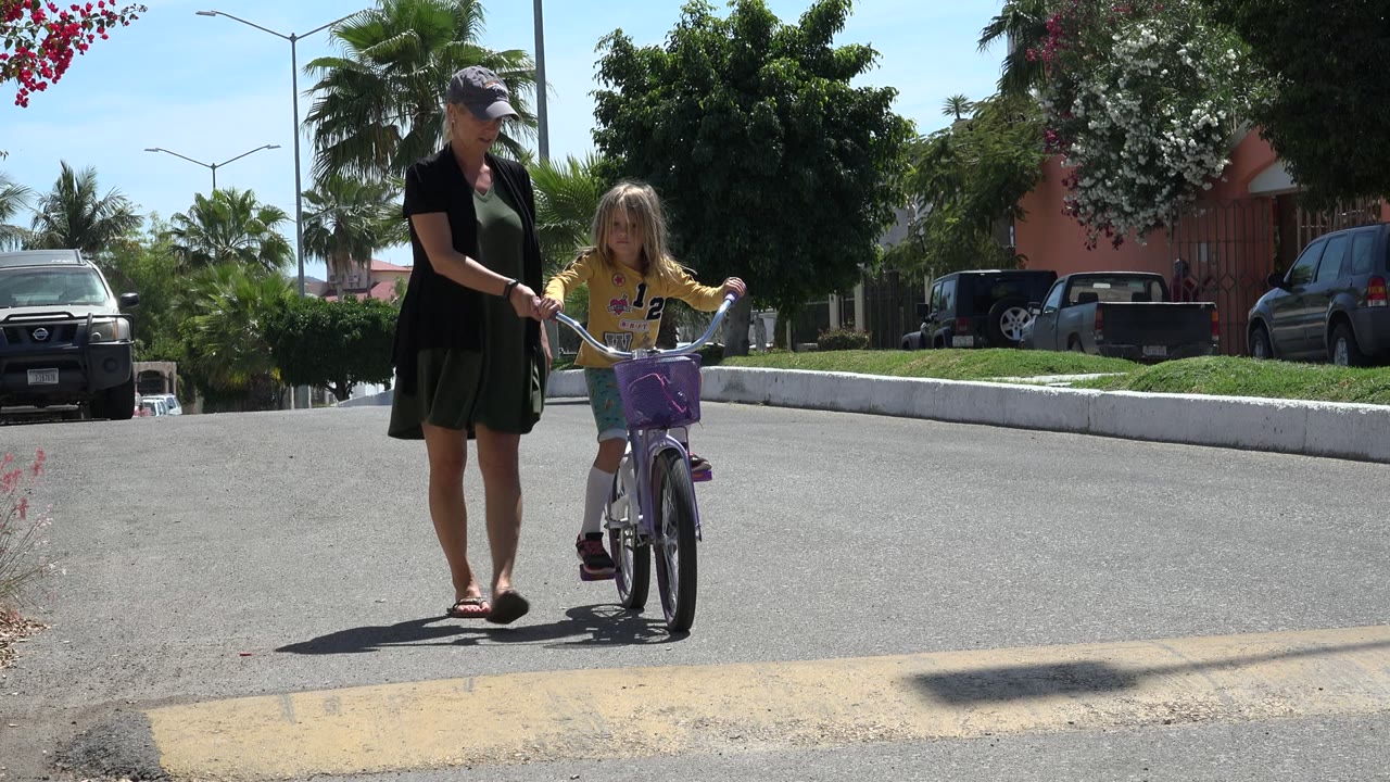 Mother walking beside a young girl on a purple bicycle in a sunny street.