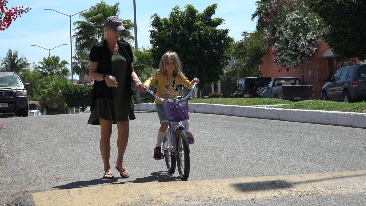 In Los Cabos, Mexico, the girl pedals forward on the purple bike while her mother walks at her left side near parked cars and palms.