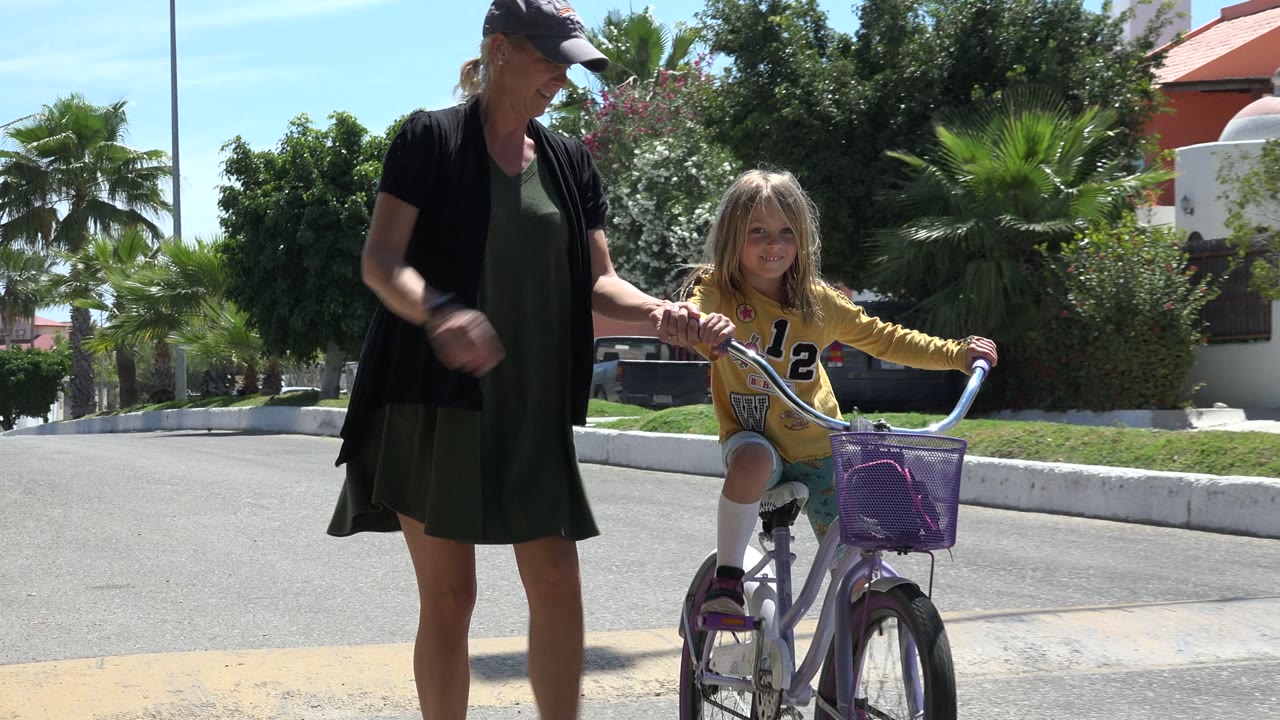 In Los Cabos, Mexico, a woman in a dark dress stands beside a young girl seated on a purple bicycle with a basket on a sunlit residential street.