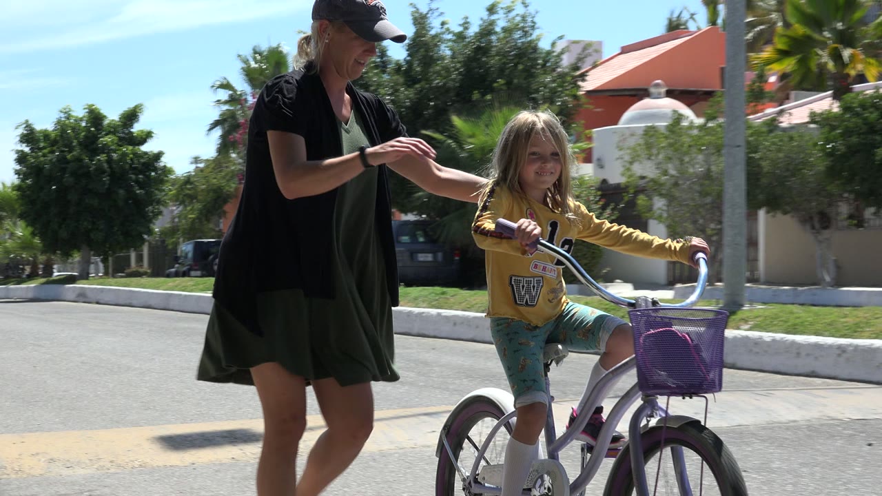 In Los Cabos, Mexico, the adult reaches toward the young girl as she holds the handlebars of a purple bicycle, with trees, pavement, and homes behind them.
