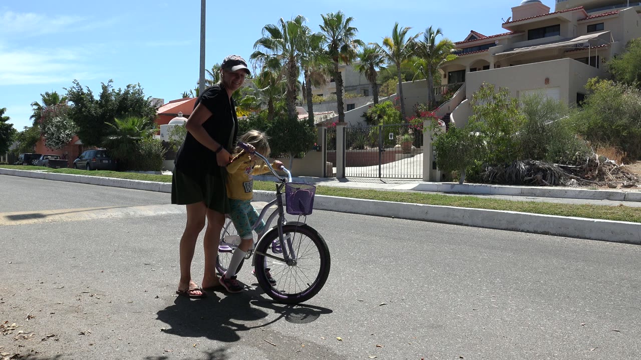 A woman stands over a child with one hand near the handlebars of a purple bicycle, with palm trees and homes behind them in Los Cabos, Mexico.