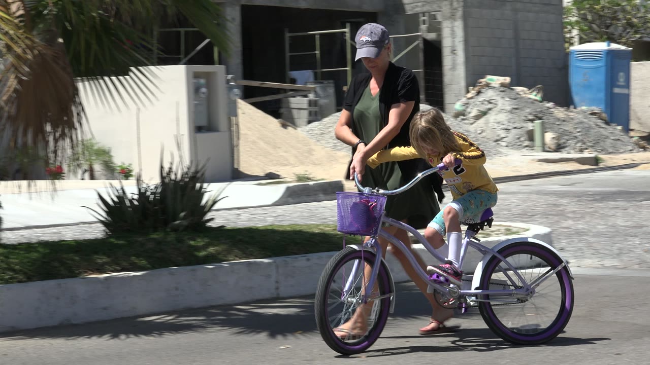 The woman leans toward the handlebars as the child sits on the small purple bike in a sunny Los Cabos street scene.