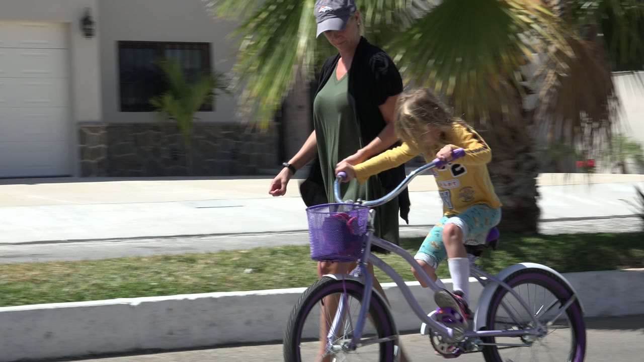 At the start of the Los Cabos clip, an adult woman walks beside a young girl riding a purple bicycle past grass, a palm trunk, and white garage doors.