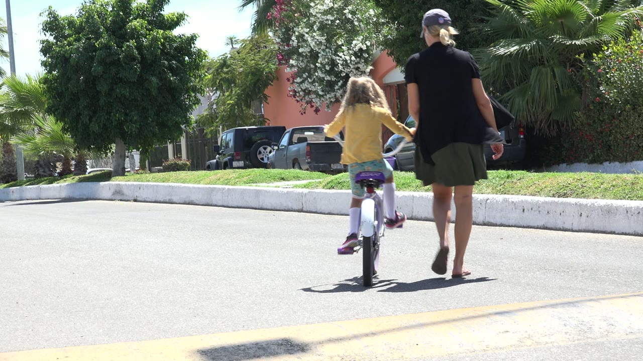 Woman walking beside a girl riding a small bicycle on a sunny street.