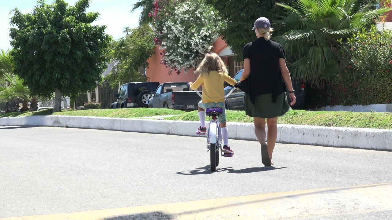 In Los Cabos, the girl pedals slightly ahead while the woman keeps pace beside her on a quiet street lined with greenery and parked cars.