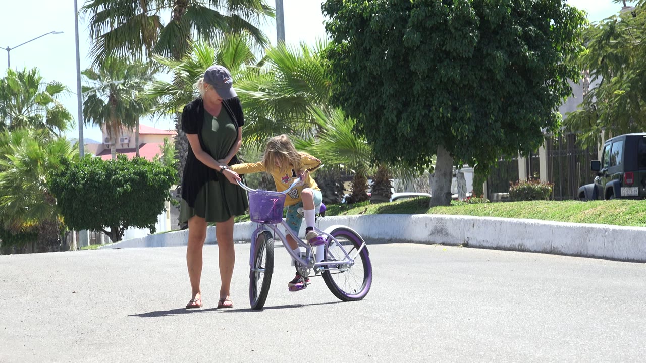 The woman leans toward the handlebars as the child looks down at the bicycle, with bright pavement and palm landscaping behind them in Los Cabos, Mexico.
