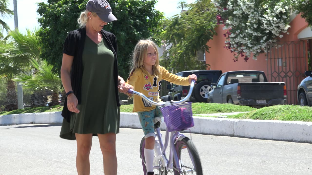 In Los Cabos, Mexico, a woman in a green dress walks beside a blonde girl starting along the street on a small white bicycle with a purple front basket.