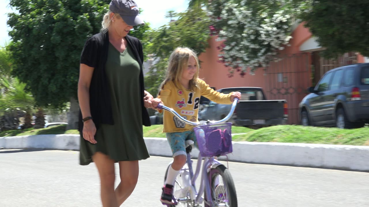 The girl pedals forward with one hand near the handlebar curve while the woman stays close beside her, with parked cars and greenery behind them in Los Cabos, Mexico.