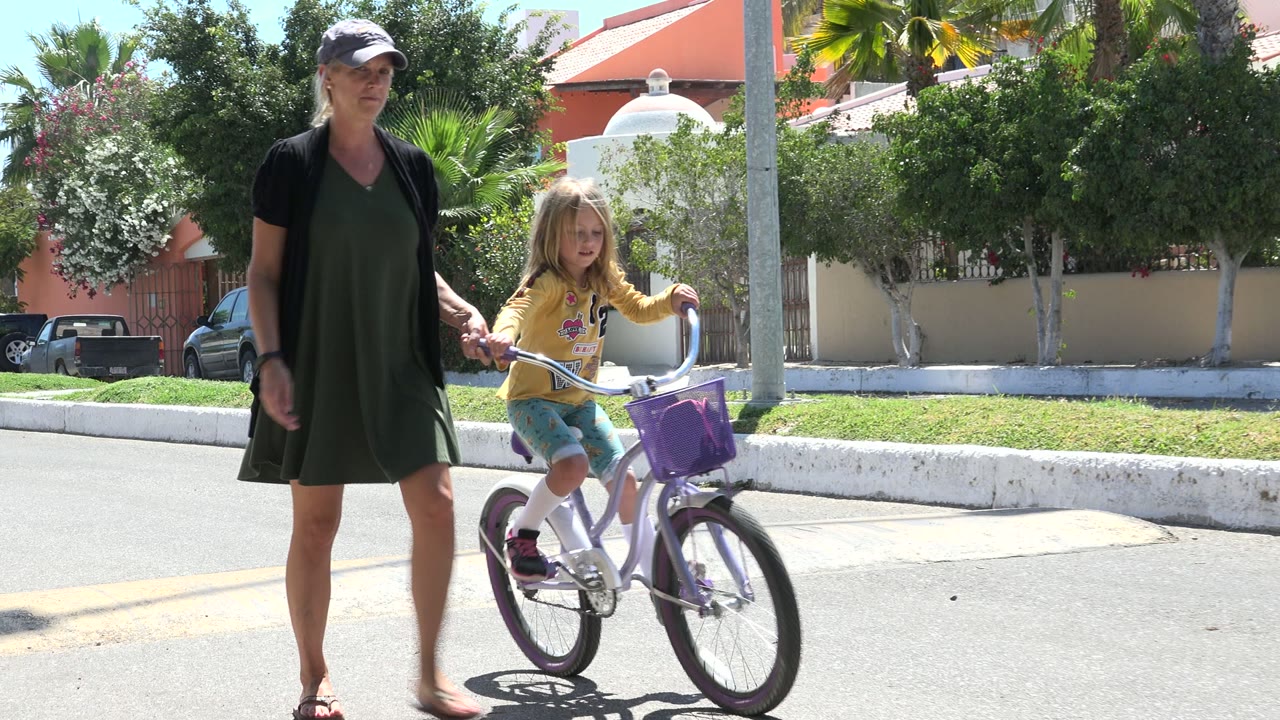 The mother walks near the child as the girl rides the small bicycle forward on a sunlit Los Cabos residential street.