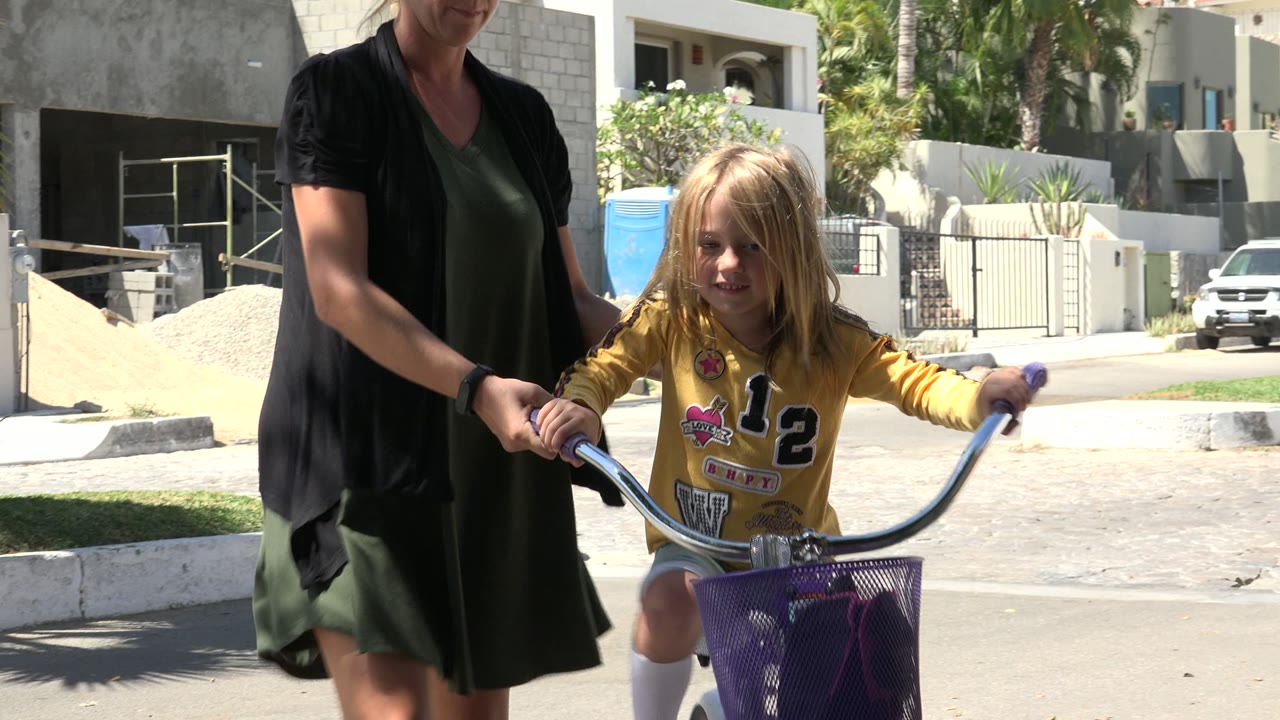 The girl rides toward the camera while the adult stays close at her side, with a purple basket, curved chrome handlebars, and bright pavement visible in Los Cabos, Mexico.