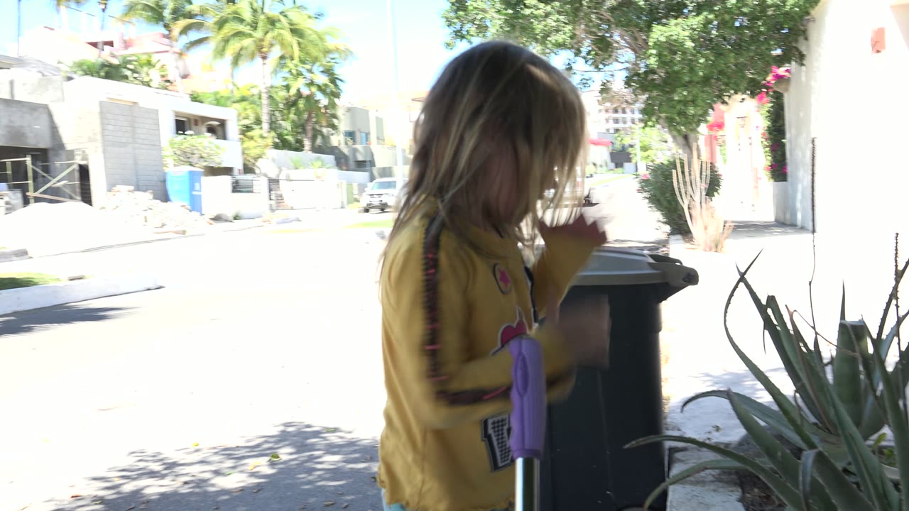 In Los Cabos, Mexico, the child stands partly turned away beside a black bin and pointed plants, with bright street and low buildings in the background.