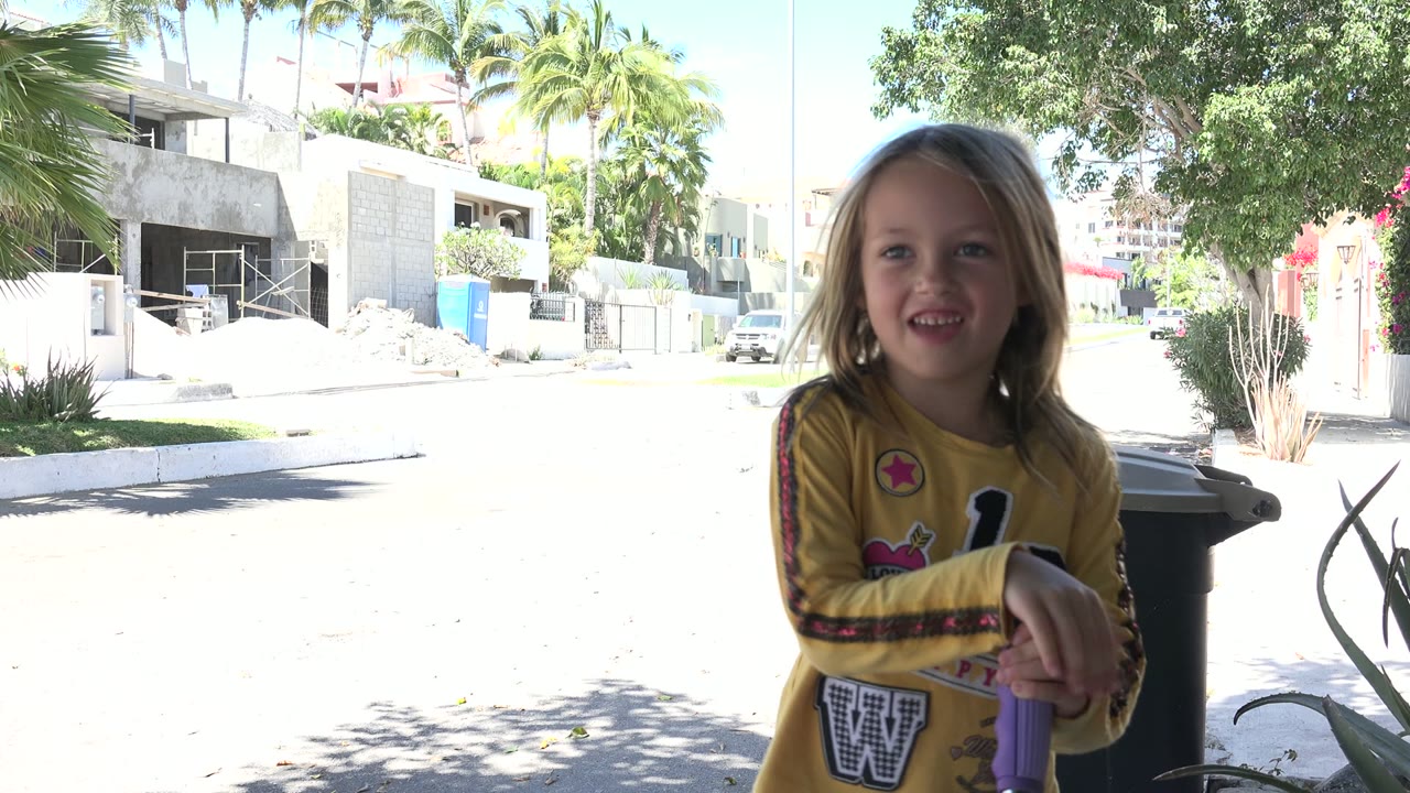 Blonde child in a yellow top faces the camera beside a purple toy on a shaded sidewalk in Los Cabos, Mexico, with bright homes and palm trees behind.