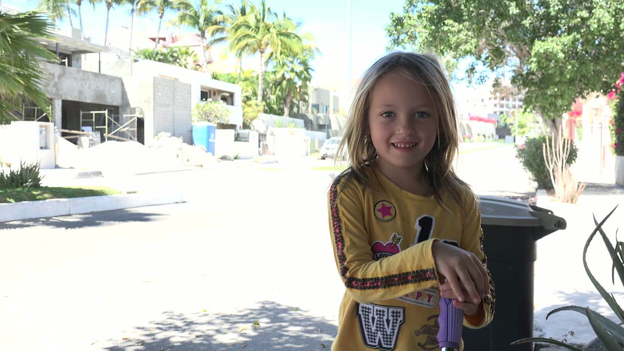 The child smiles near the purple toy handle while the shaded foreground contrasts with a bright residential street in Los Cabos, Mexico.