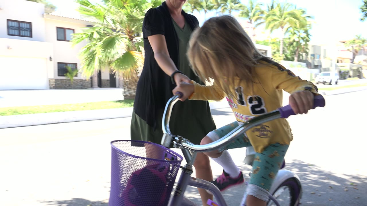 The child bends toward the handlebars of the bicycle while the adult keeps one hand near the front of the bike in the Los Cabos street setting.