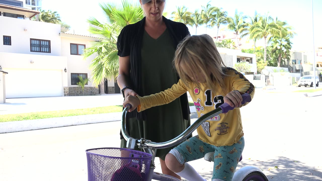 In Los Cabos, Mexico, an adult stands behind a young child leaning over a small bicycle with purple grips and basket near a bright residential street.