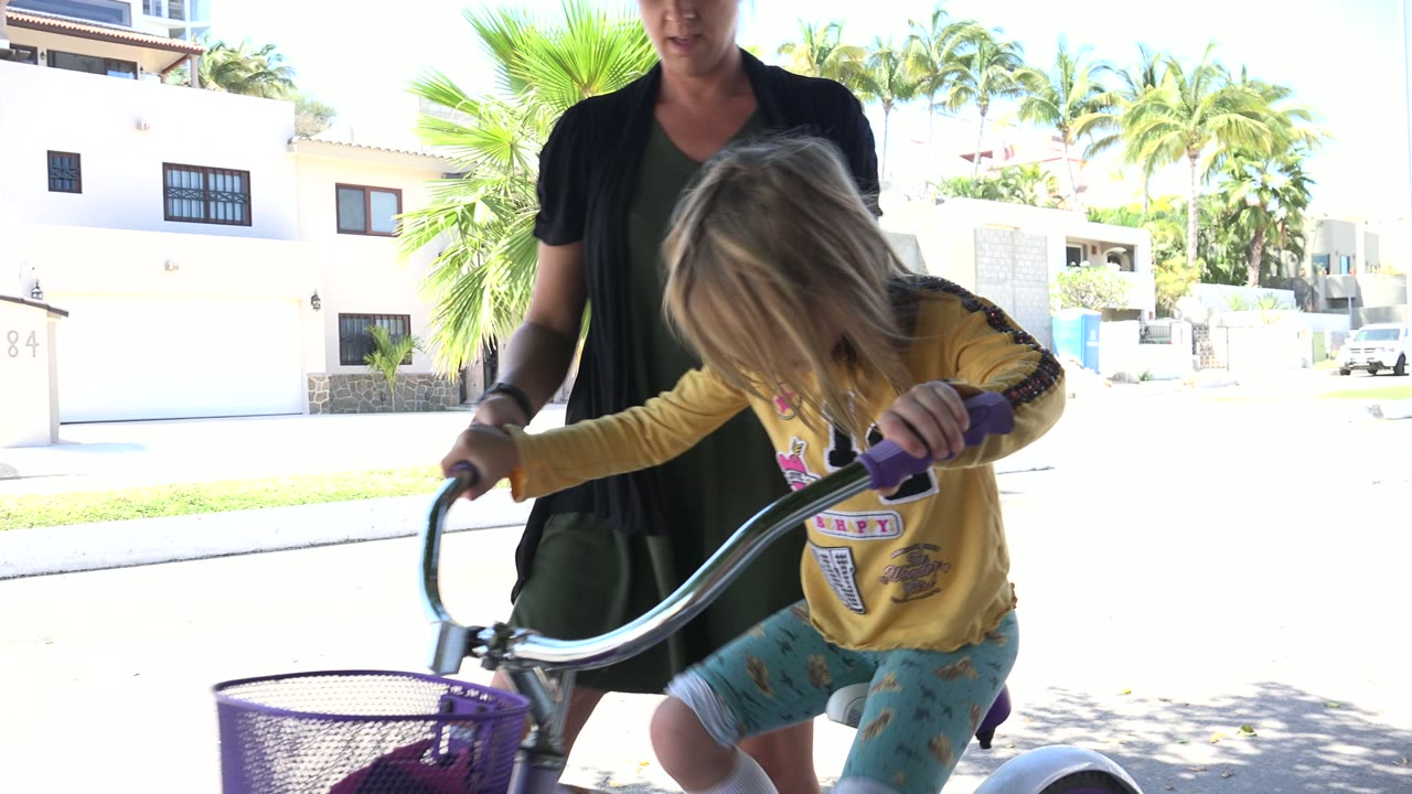 The child bends toward the handlebars of the small bicycle as the adult remains partly cropped behind them on a sunlit Los Cabos street.