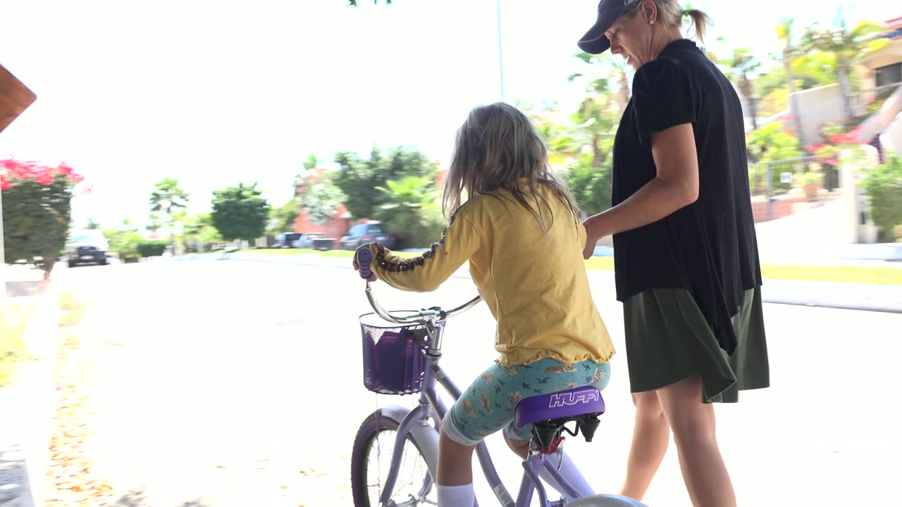 At the start of the Los Cabos clip, a young girl in a yellow top sits on a purple bicycle while an adult woman stands close beside her on a sunlit street.