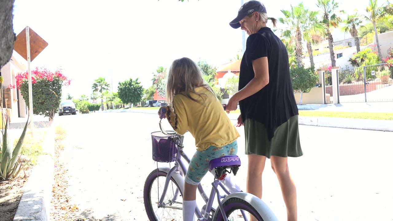 One second in, the girl leans over the handlebars of the purple bicycle as the woman watches beside her in the bright Los Cabos street scene.