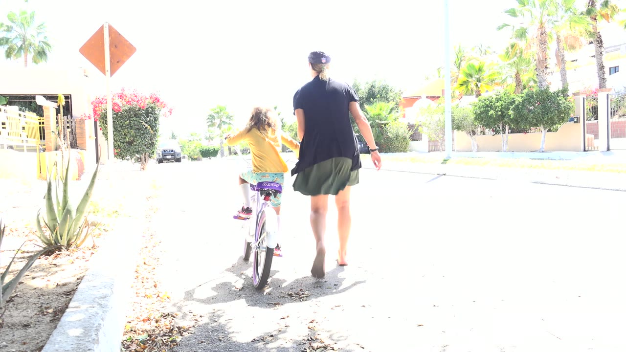 In Los Cabos, Mexico, a woman walks close behind a young girl riding a small bicycle along a sunlit residential street with palms and desert plants nearby.