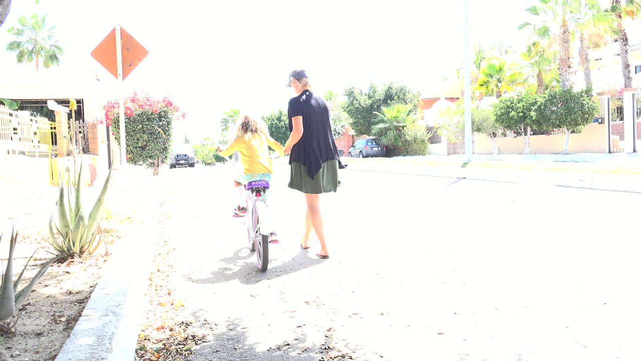 The girl pedals slightly farther ahead while the woman walks beside her on the overexposed street, with a curb, agave-like plants, palms, and homes in view in Los Cabos, Mexico.