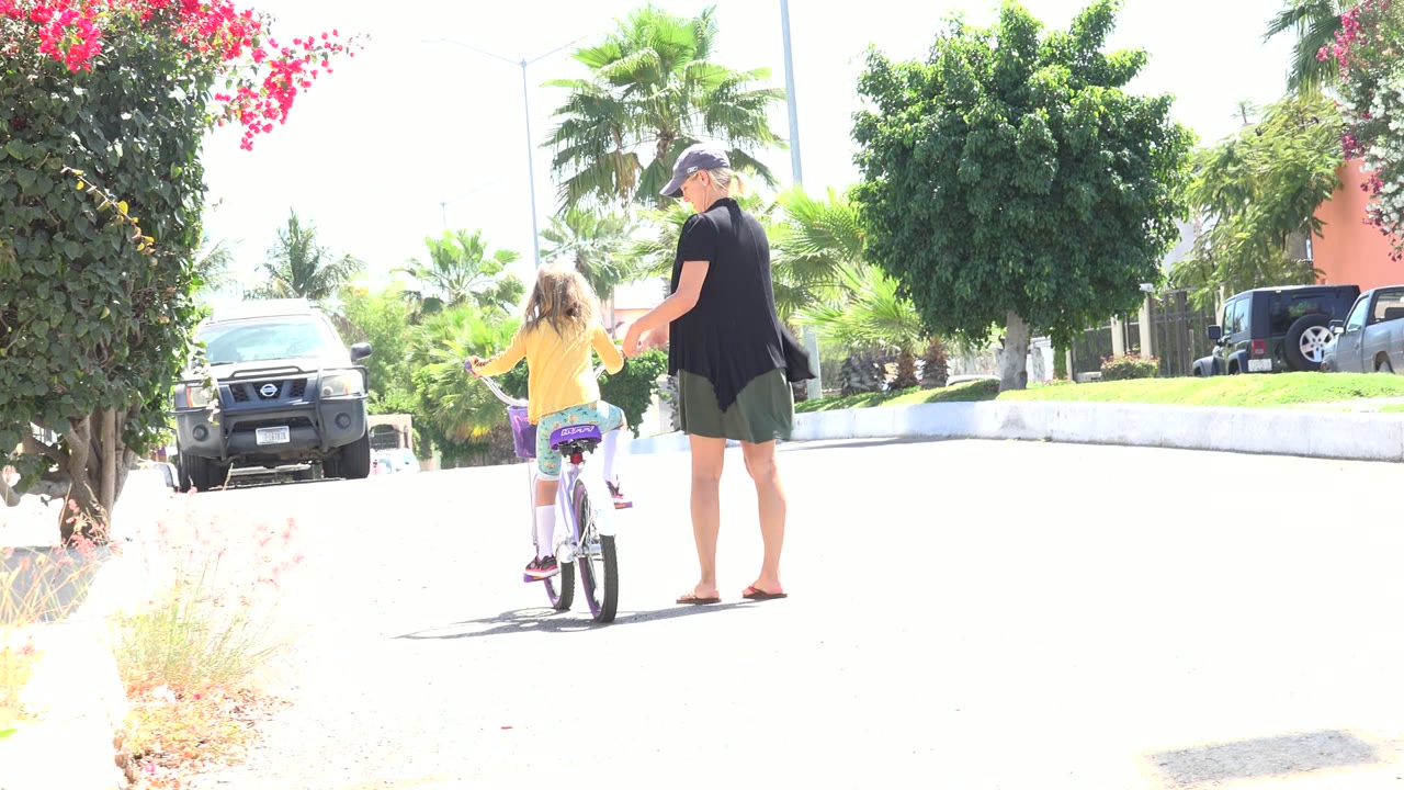 The child pedals farther along the Los Cabos street while the adult woman keeps one hand near the bicycle for balance.