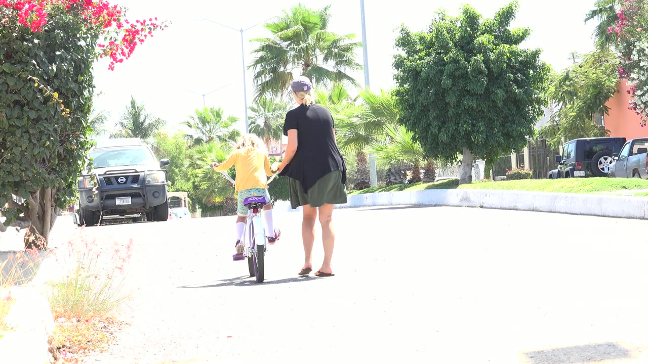 Woman walking beside a child riding a bicycle on a bright street.