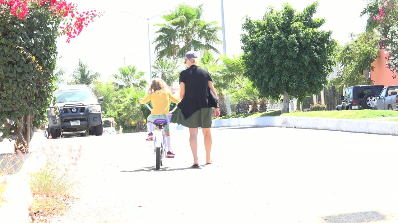 The girl rides farther down the Los Cabos street while the woman stays at her right side, with a parked vehicle on the left and palms and shrubs in bright sun behind them.