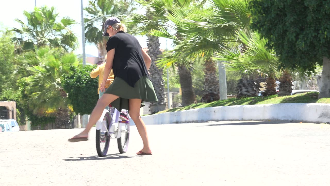 At 0:01 in Los Cabos, Mexico, the woman steps alongside the child's bicycle as the pair move across sunlit pavement with palms and shrubs behind them.