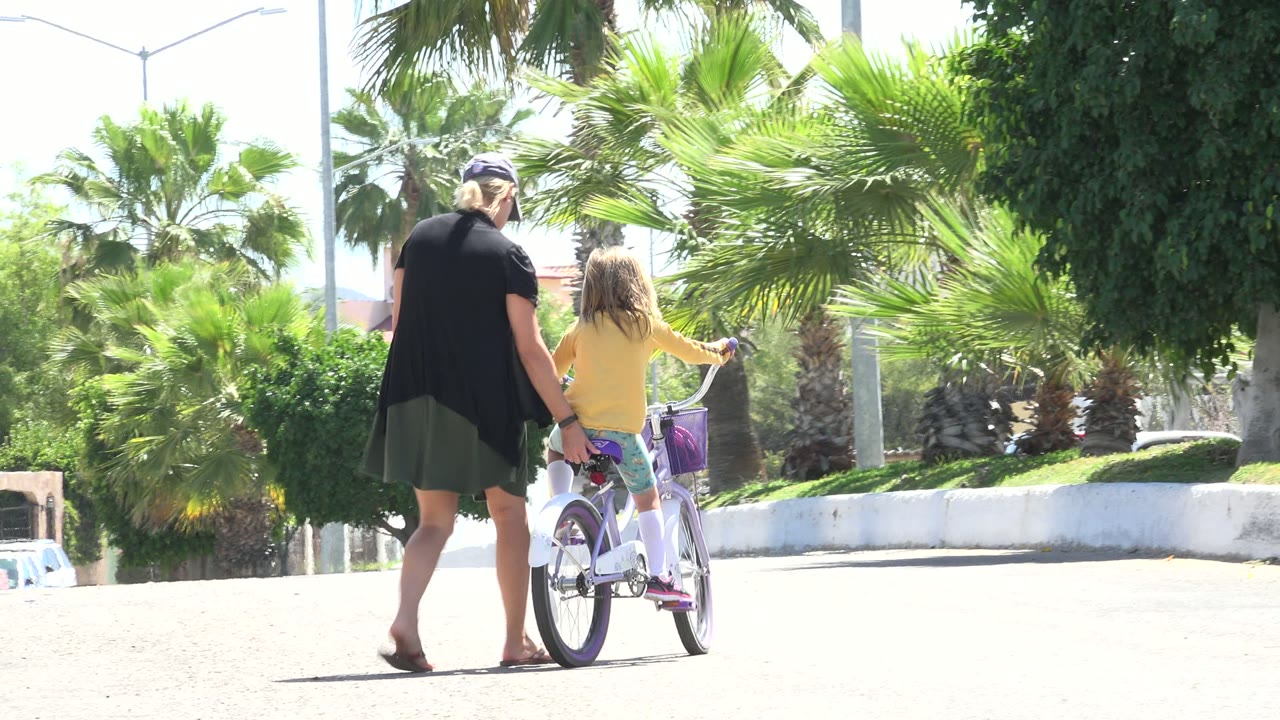In Los Cabos, Mexico, the child rides slightly farther ahead while the adult walks close behind, with bright pavement, palm fronds, shrubs, and a white retaining edge visible.