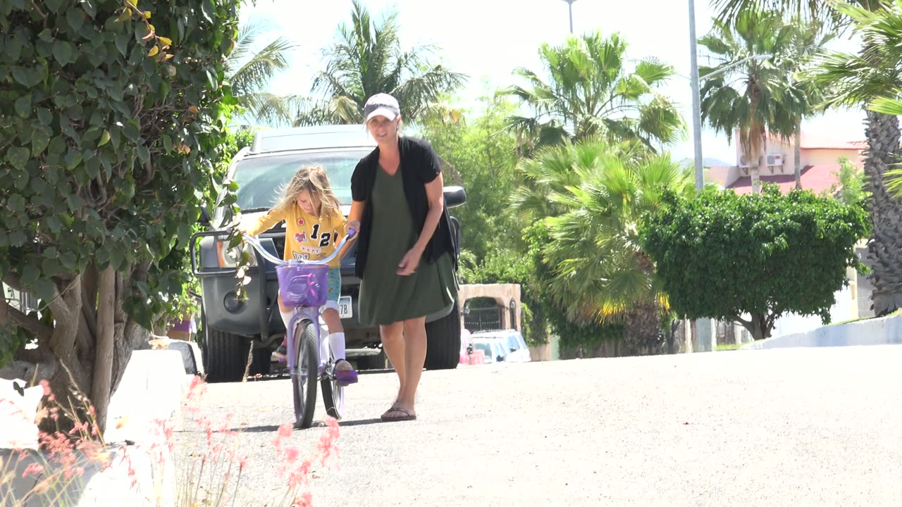 Girl riding a purple bicycle beside a woman on a sunny street.