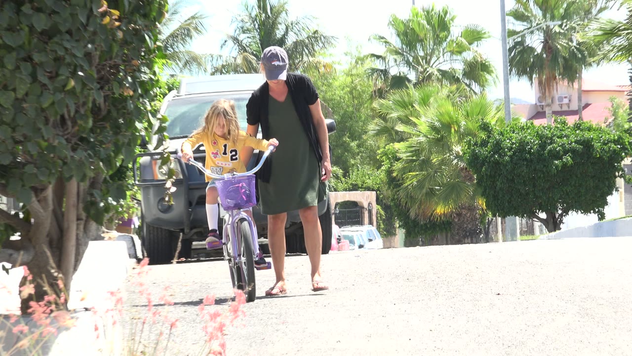 The girl pedals forward on the purple bicycle while the woman walks next to her, with palms and a dark parked vehicle behind them in Los Cabos.