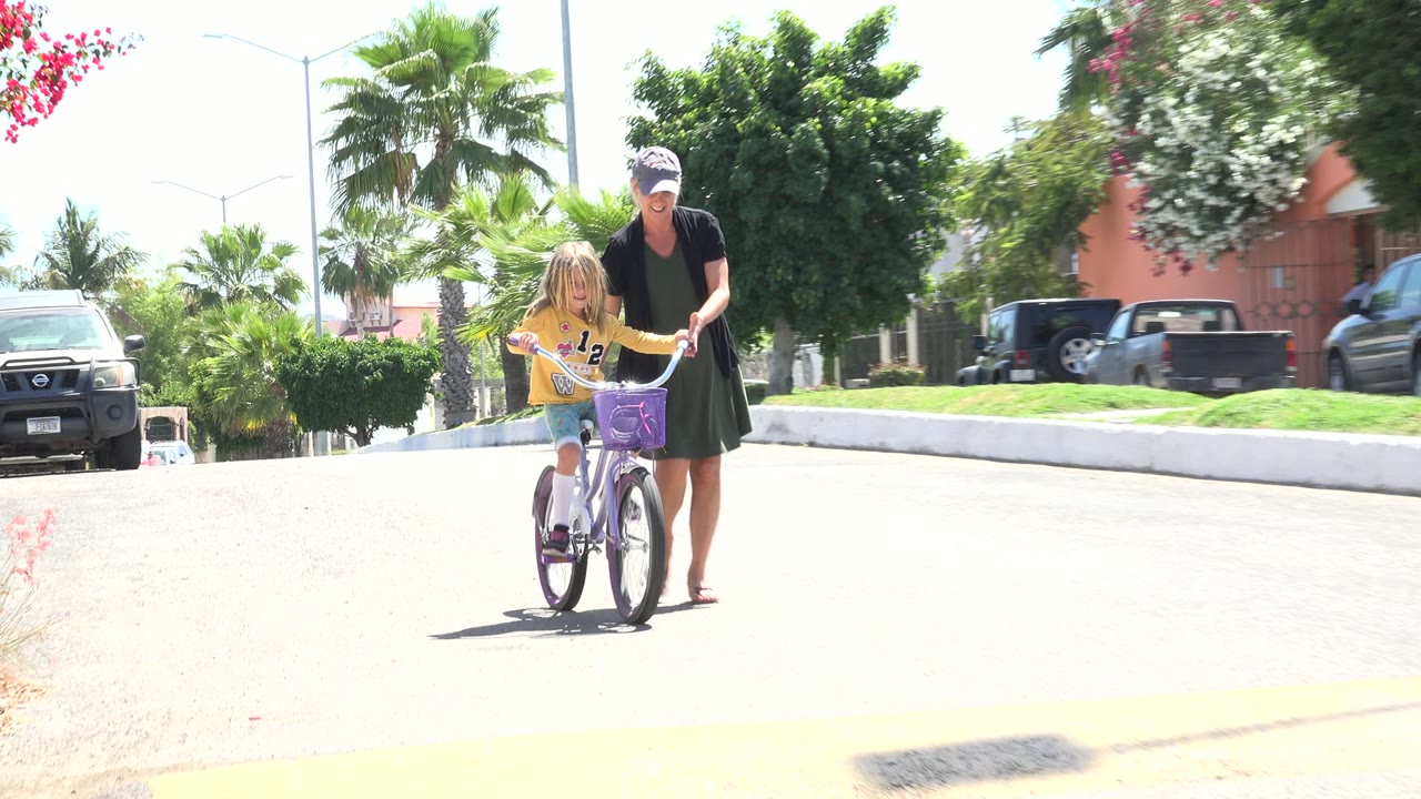 Woman steadies a child on a small bicycle in a sunny residential street.