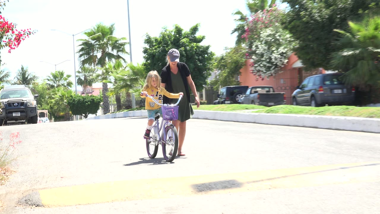 The girl pedals slightly farther into the Los Cabos residential street while the woman stays next to the bicycle handlebars, with parked vehicles and palms behind them.