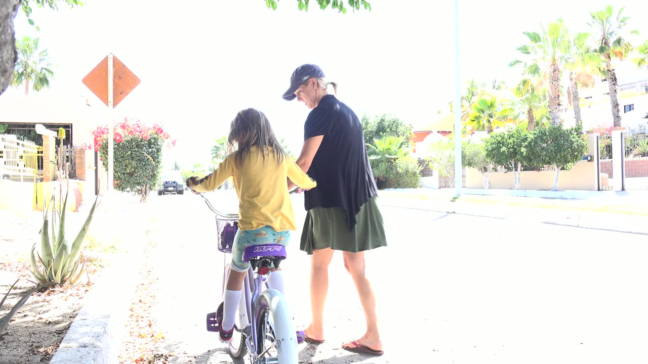 Child on a bicycle with an adult woman walking beside her on a bright road.
