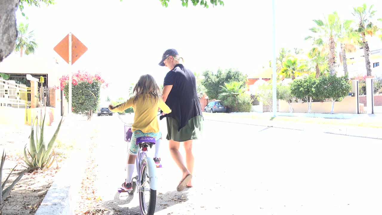 The child moves farther down the Los Cabos road on the bicycle while the adult woman walks alongside, with palms, shrubs, and parked cars in the bright background.