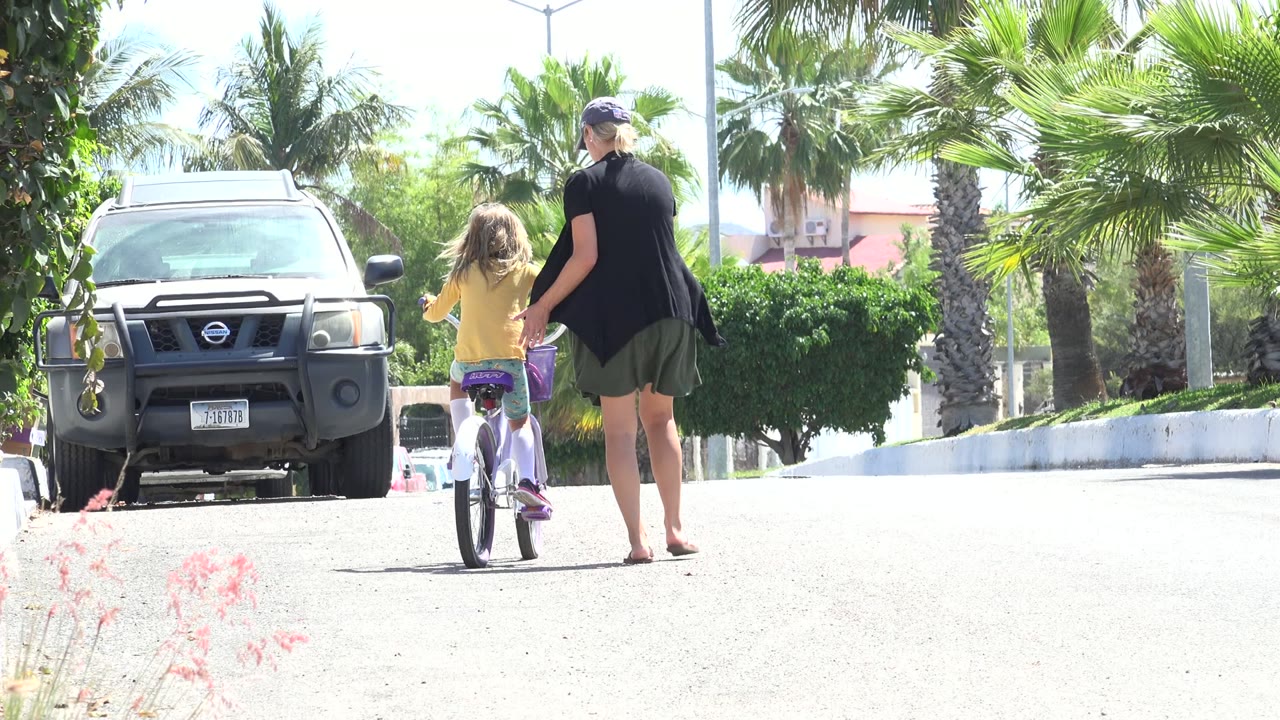 In Los Cabos, Mexico, an adult woman walks beside a child on a small bicycle near a parked Nissan vehicle and palm trees at the start of the clip.